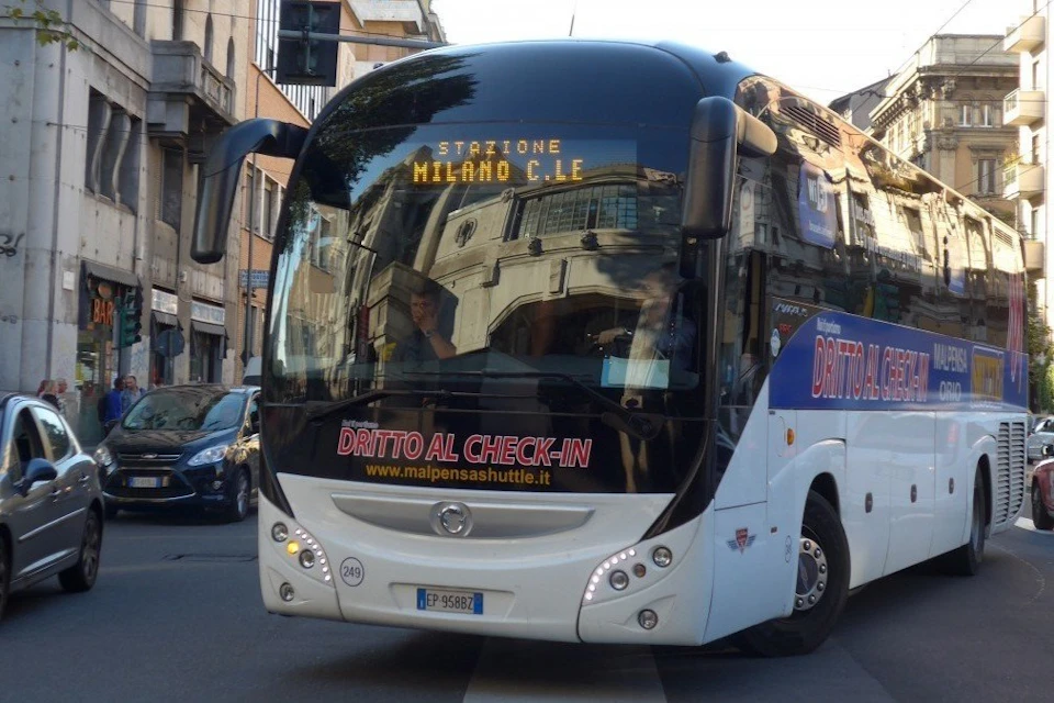 A shuttle bus labeled "Malpensa Shuttle" with a display reading "Stazione Milano C.le" driving on a city street.