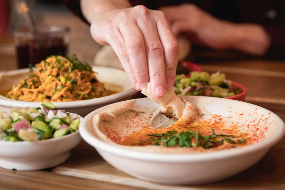 A hand dipping bread into a bowl of hummus, accompanied by plates of salad and vegetables on a wooden table.