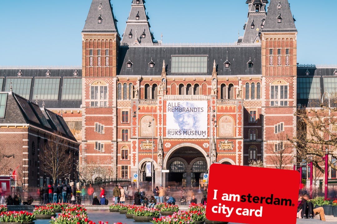 Rijksmuseum i Amsterdam en solig dag, med tulpanblommor i förgrunden och människor som går i närheten.