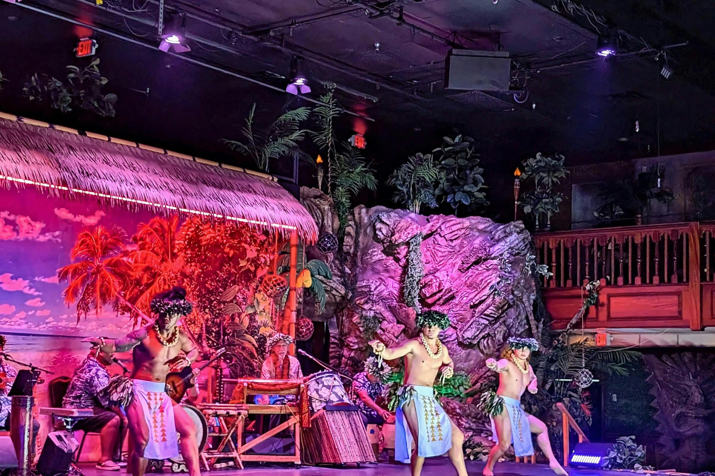 Performers in traditional Polynesian attire dancing on a themed stage with tropical and rock decorations.