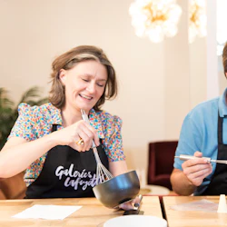Två leende vuxna i förkläden som vispar ingredienser i skålar på en matlagningskurs.