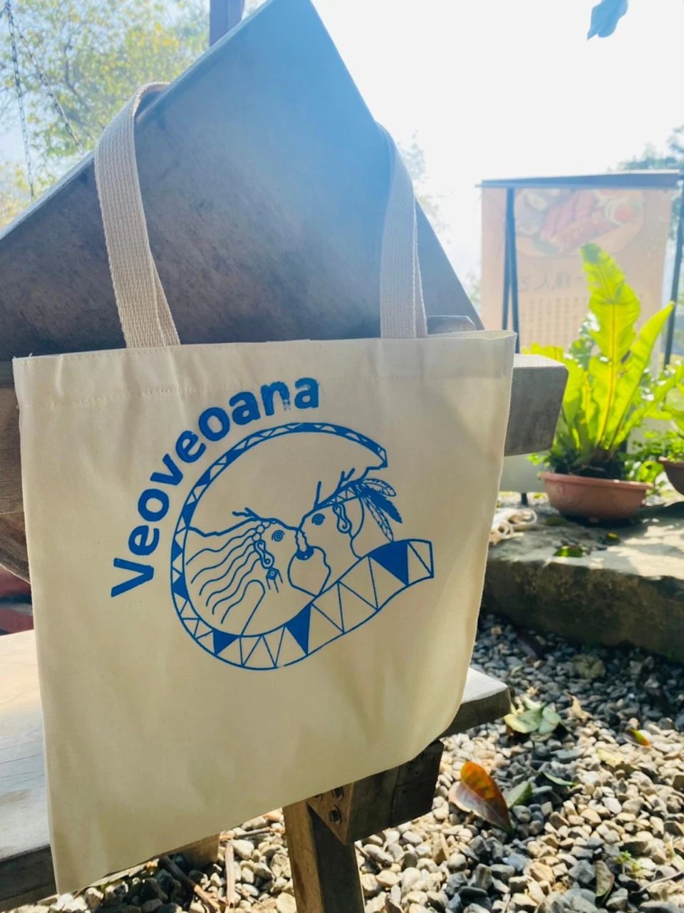 A beige tote bag with the text 'Veoveoana' and a blue design of two faces, placed outdoors on wooden furniture near plants.