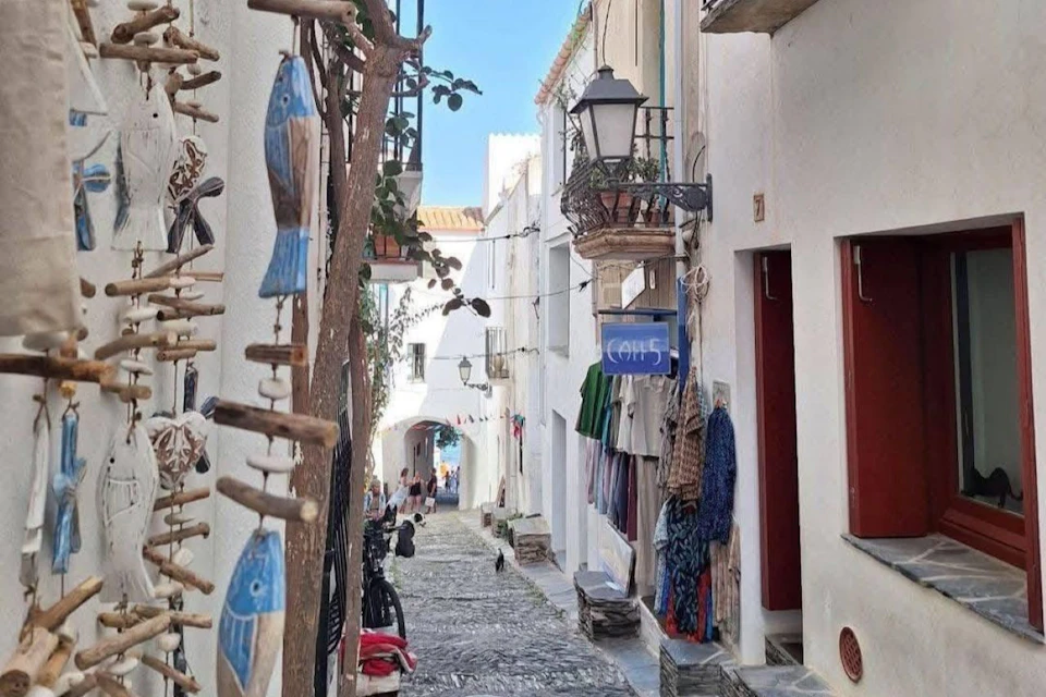 A narrow cobbled alley in a Mediterranean village, adorned with hanging decorations, plants and colourful textiles.