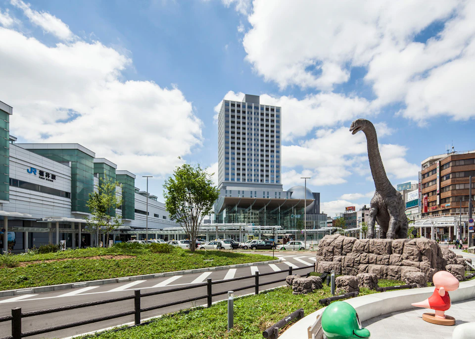 Cityscape with a modern tall building, a dinosaur statue in a park area, and a clear blue sky with clouds.