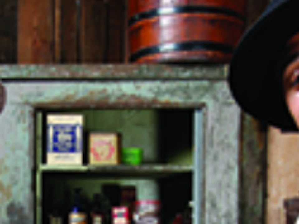 Two people dressed in vintage attire stand in front of a wooden shelf stocked with various jars and boxes.