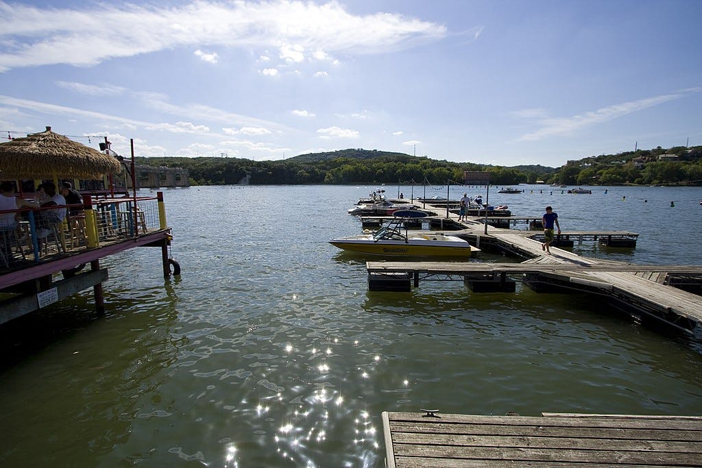Mensen dineren onder een rieten dak aan een meer met boten aangemeerd aan houten pieren en beboste heuvels op de achtergrond.