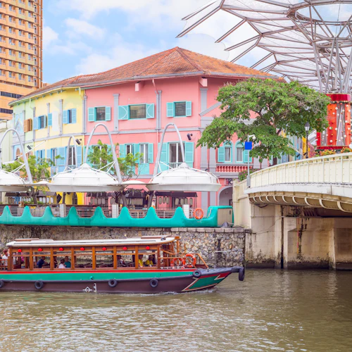 Bunte Gebäude mit blauen Fensterläden säumen eine Uferpromenade unter einem klaren Himmel. Ein Boot mit Passagieren fährt auf dem Wasser.