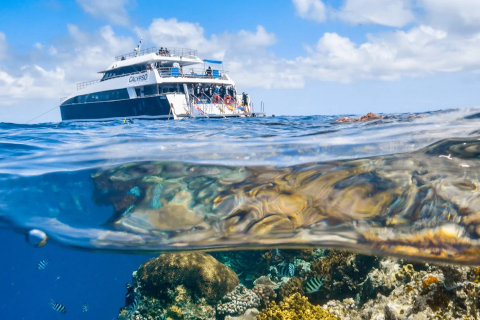 Partially submerged view of a boat named "Calypso" with people onboard and visible underwater coral reef. Clear sky background.