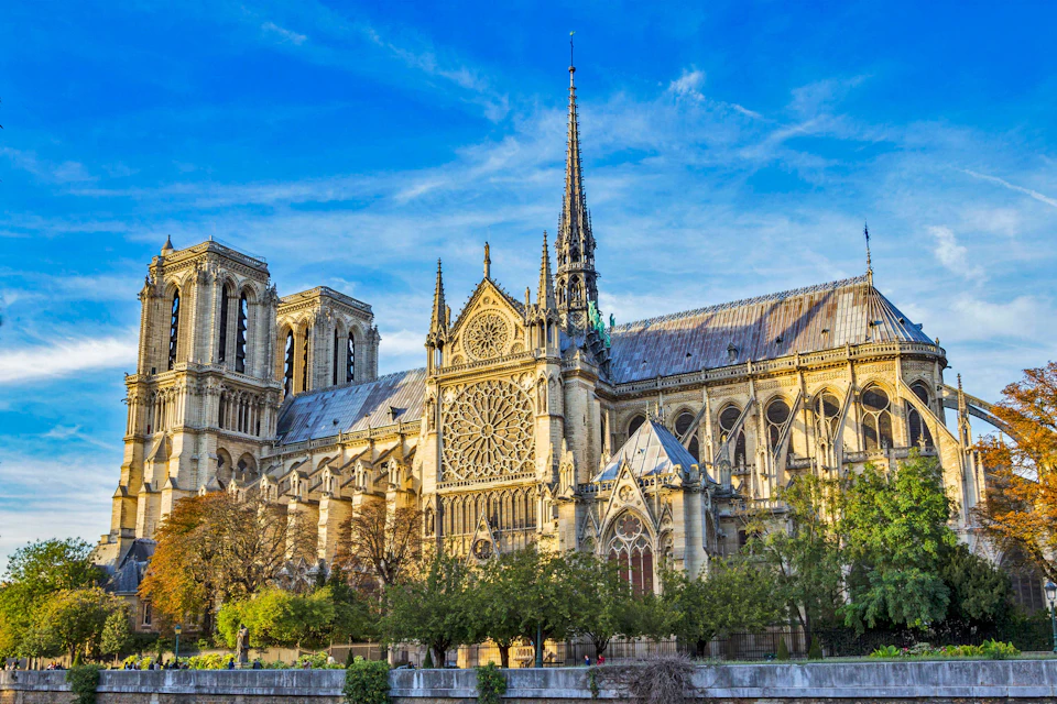 Gothic cathedral with large rose window, tall spire, and flying buttresses against a bright blue sky with some greenery in the foreground.