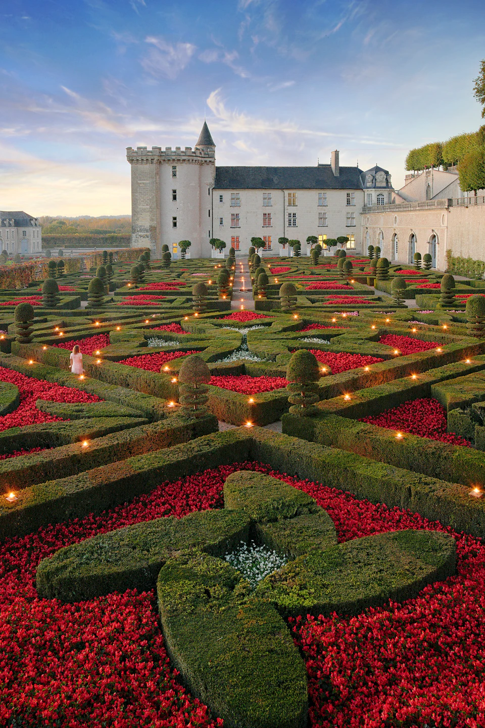 Intricate formal garden with manicured hedges, vibrant red flowers, and a historic castle in the background at sunset.
