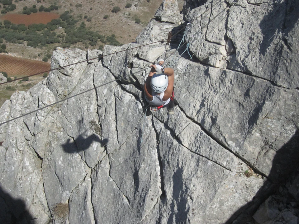 A person wearing a helmet and harness is rock climbing on a steep, rocky cliff face with a shadow cast beside them.