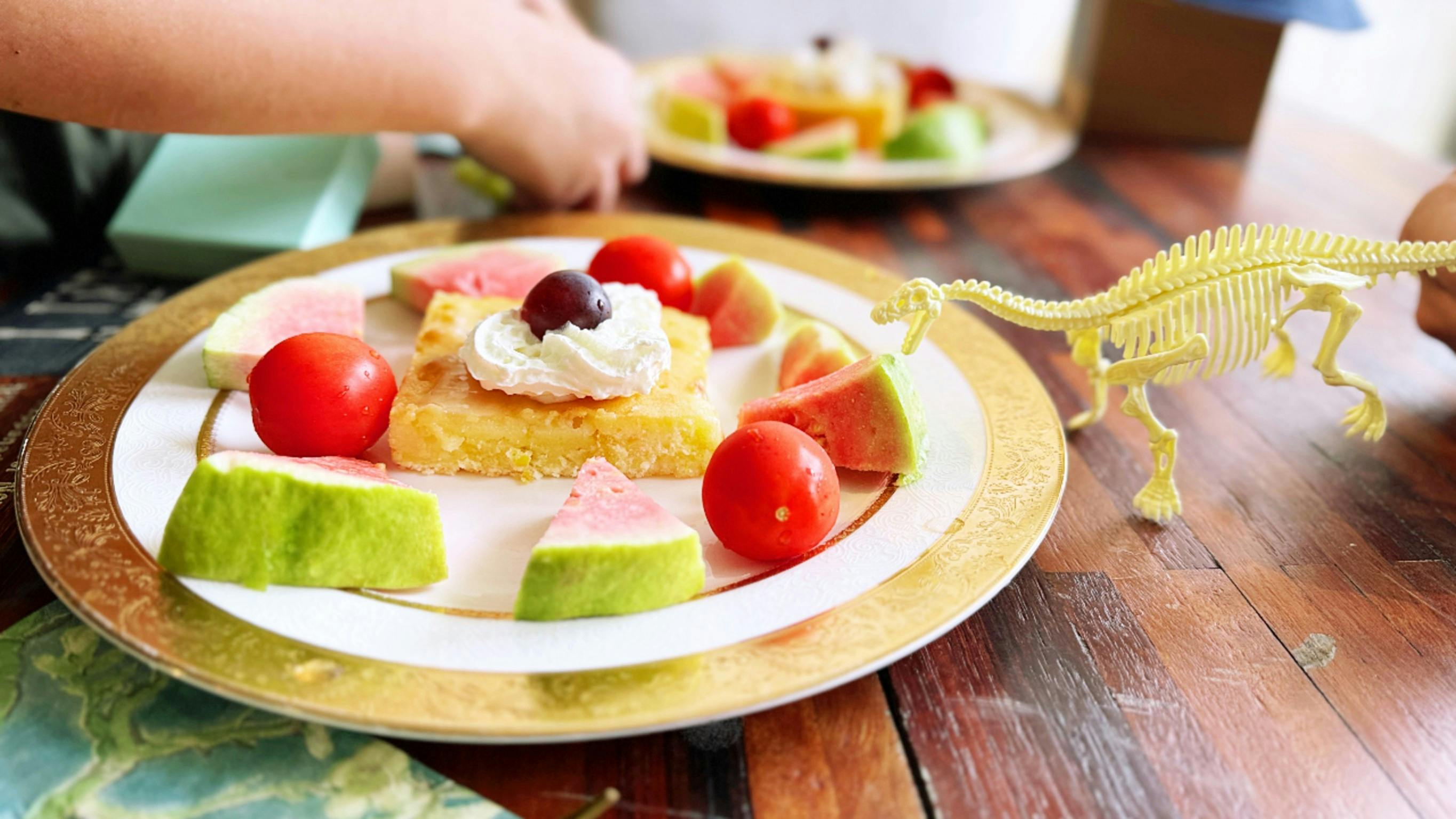A plate with a pastry topped with whipped cream and a cherry, surrounded by watermelon slices and cherry tomatoes.