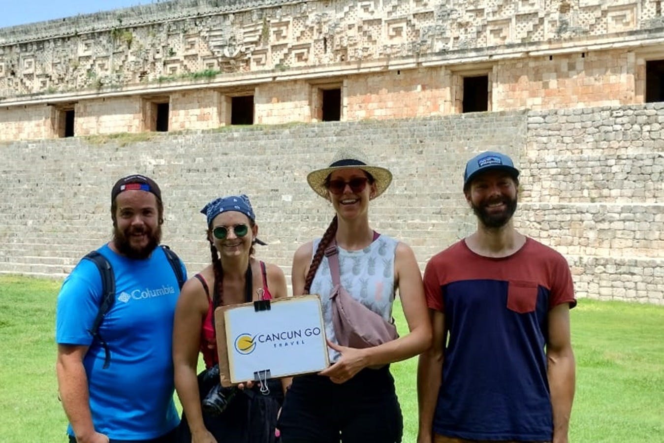 Trois personnes en tenue décontractée posant devant une ancienne structure en pierre, tenant un panneau "Cancun Go Travel".