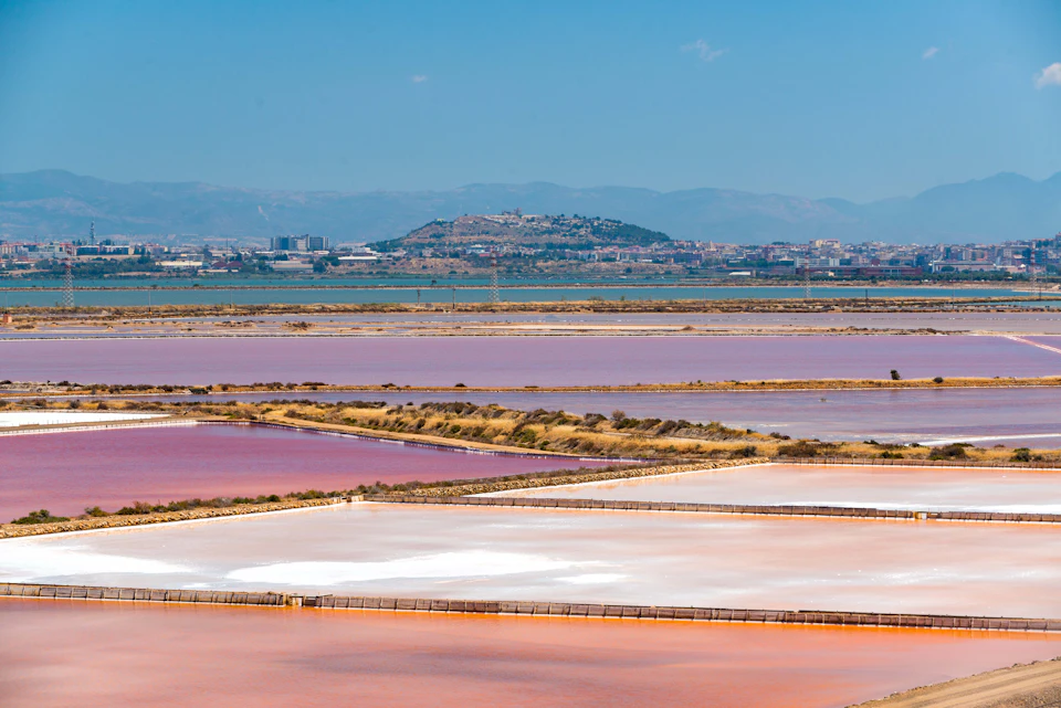 Multi-colored salt evaporation ponds with pink, purple, and white hues, situated near an urban area with hills in the background.