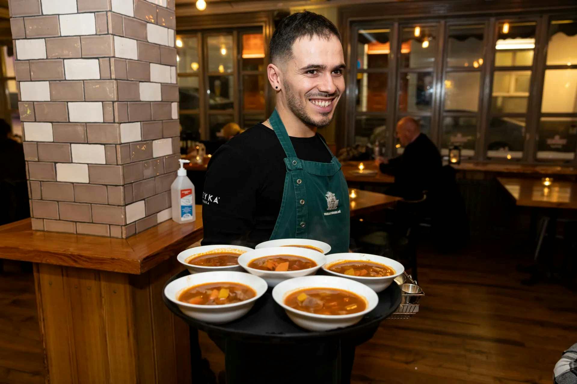 A smiling server in an apron holds a tray with six bowls of soup inside a warmly lit restaurant.