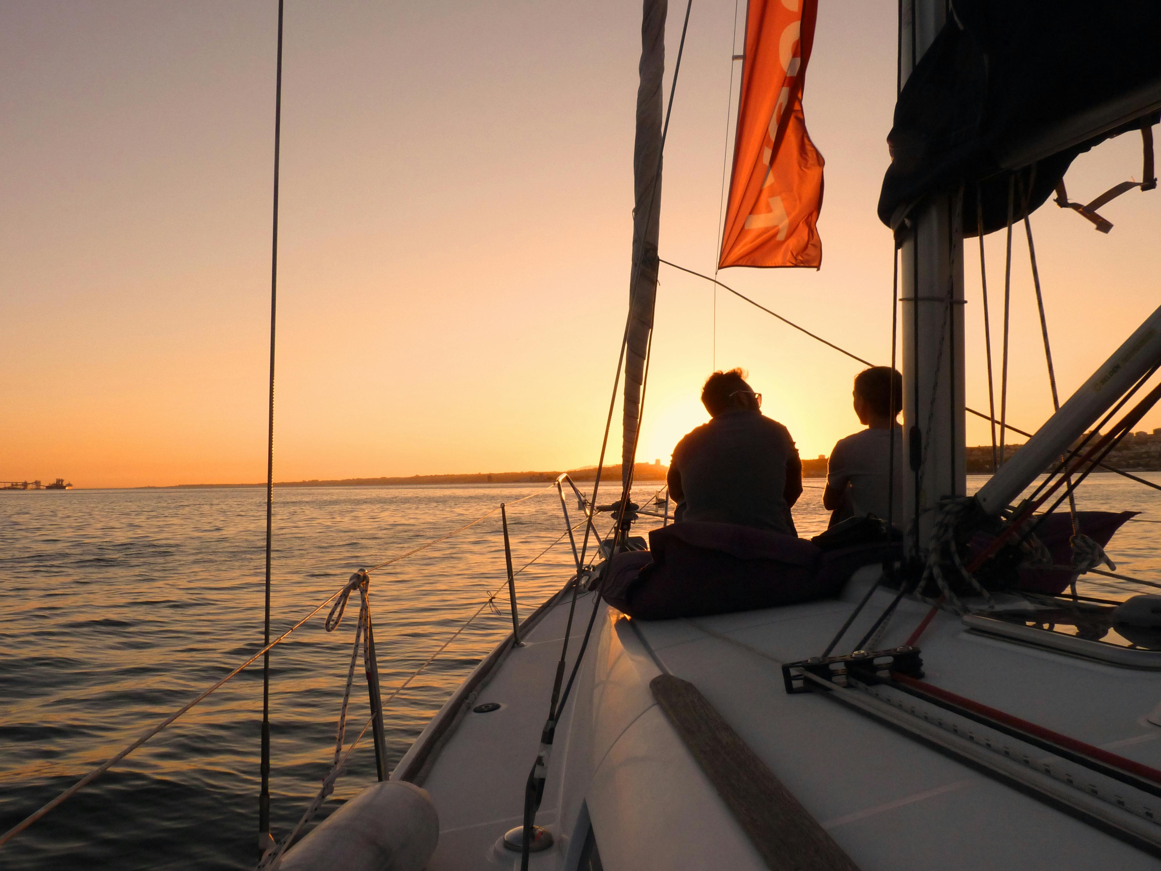 Two people sitting on a sailboat at sunset, overlooking calm sea waters and distant shoreline.