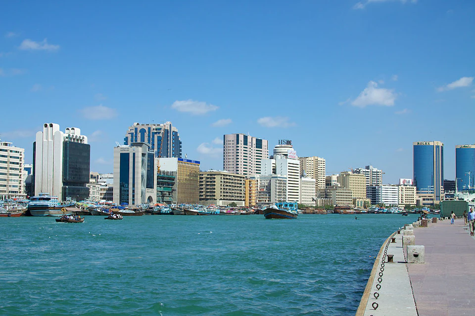 Skyline of a city with tall buildings along the waterfront, boats docked, and a few clouds in the blue sky.