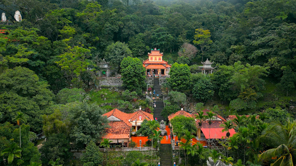 A temple complex with orange-roofed buildings and a staircase leading up, surrounded by lush green trees and forested hills.