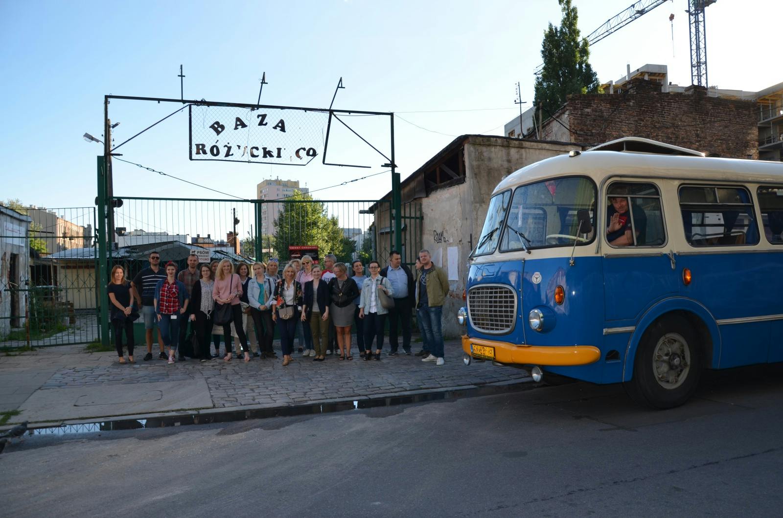 石畳の通りに青と白のバスが停まっている「Baza Różycki Co」と書かれた門の近くに、人々が立っています。