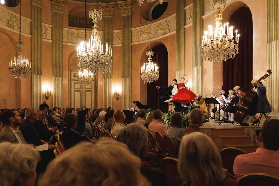 A formal ballroom with chandeliers, a live music ensemble, and a dance performance for an audience seated in rows.
