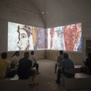 People sitting on benches in a dimly lit room, watching a large projection of ancient fresco-style artwork on two walls.