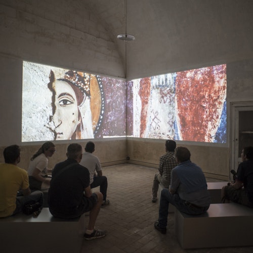 People sitting on benches in a dimly lit room, watching a large projection of ancient fresco-style artwork on two walls.