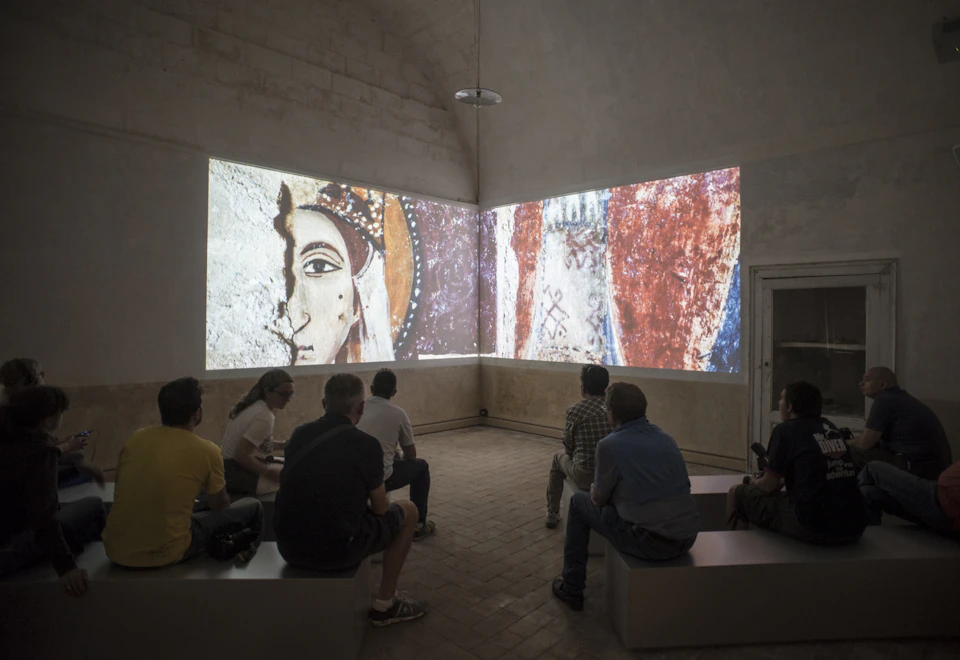 People sitting on benches in a dimly lit room, watching a large projection of ancient fresco-style artwork on two walls.