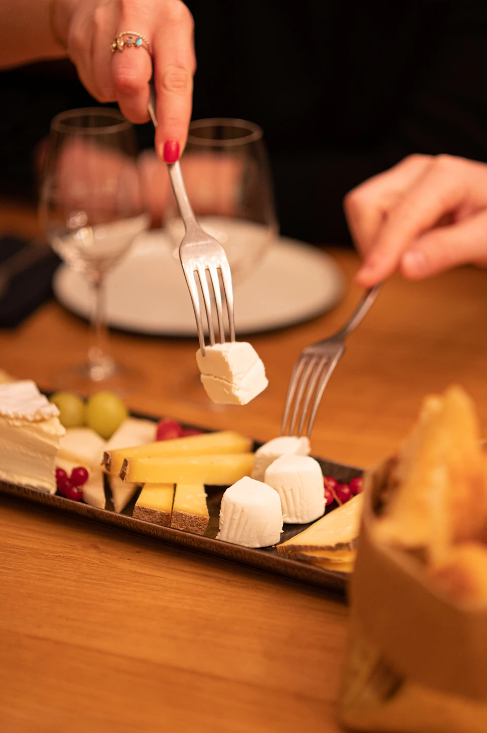 Two forks picking up pieces of cheese from a platter with various cheeses, grapes, and red berries on a wooden table.