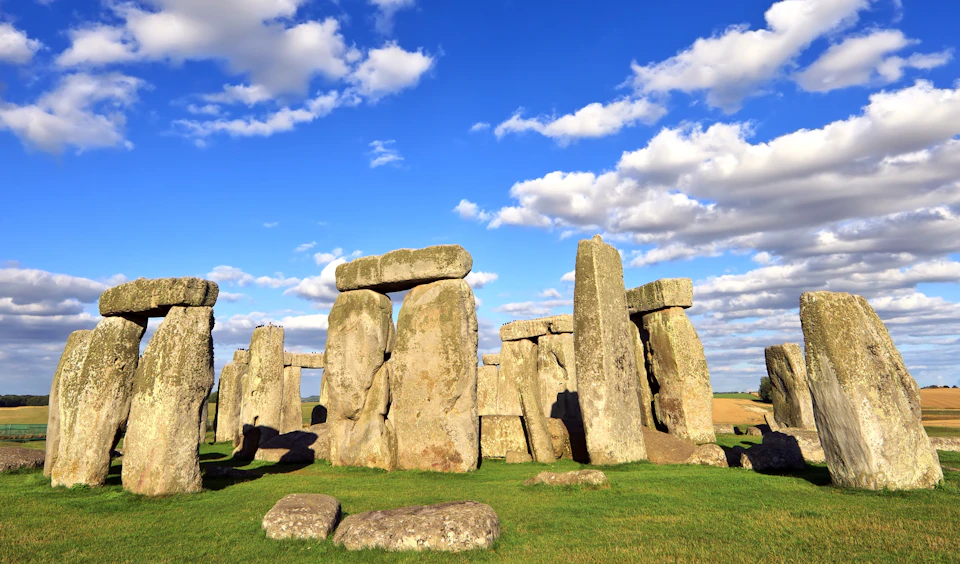 Stonehenge under a bright blue sky with scattered clouds, surrounded by green grass and rocks.