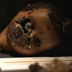 Ett skadat dockhuvud med saknade delar som visas på en yta, med ett dokument som är delvis synligt under det.