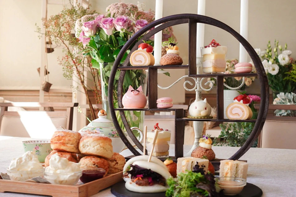 A table set with an assortment of pastries, desserts, and a steamed bun, with flowers and a decorative shelf in the background.
