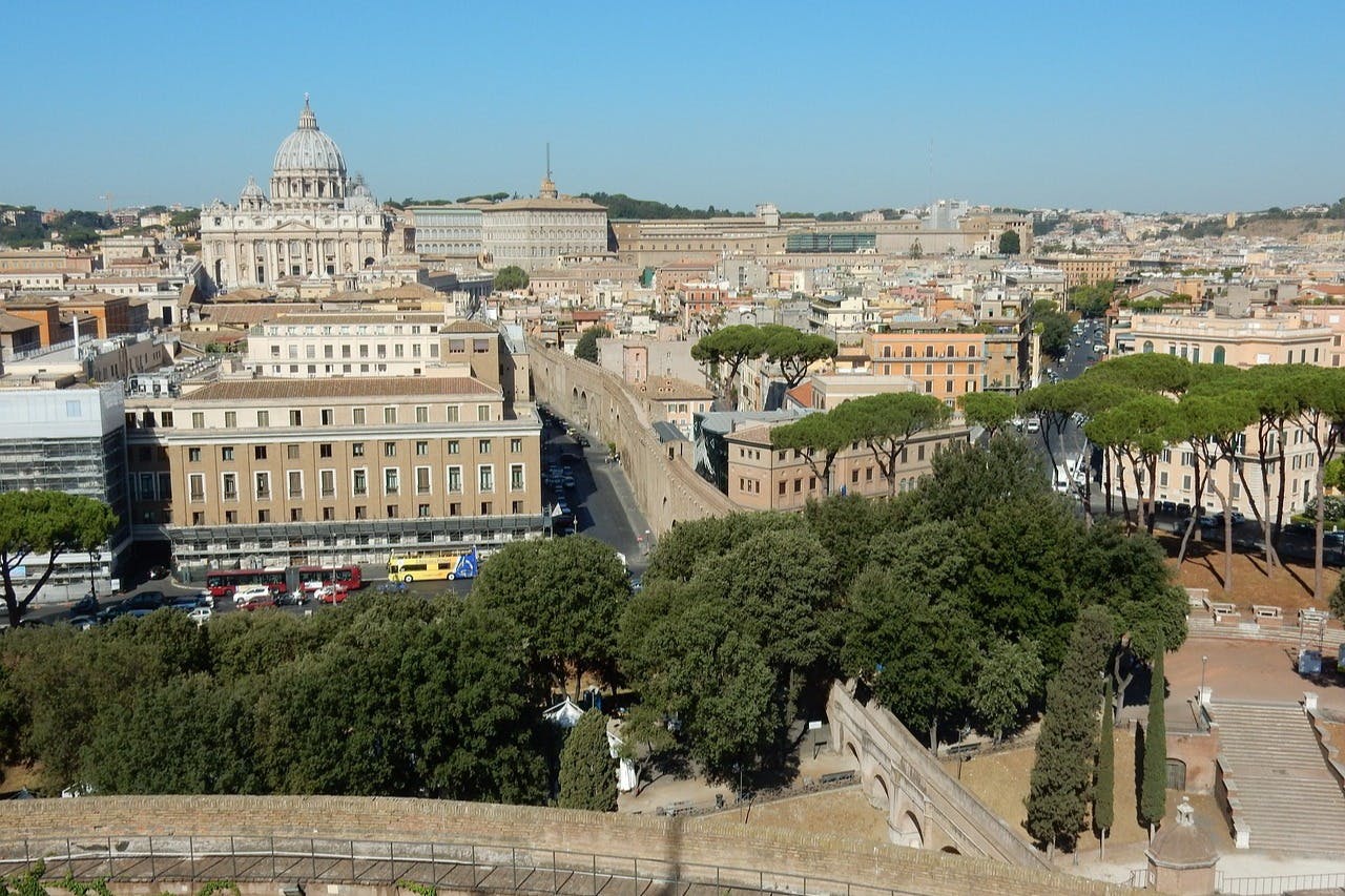 Aerial view of a cityscape with historic buildings, tree-lined streets, and the dome of a large cathedral in the background.