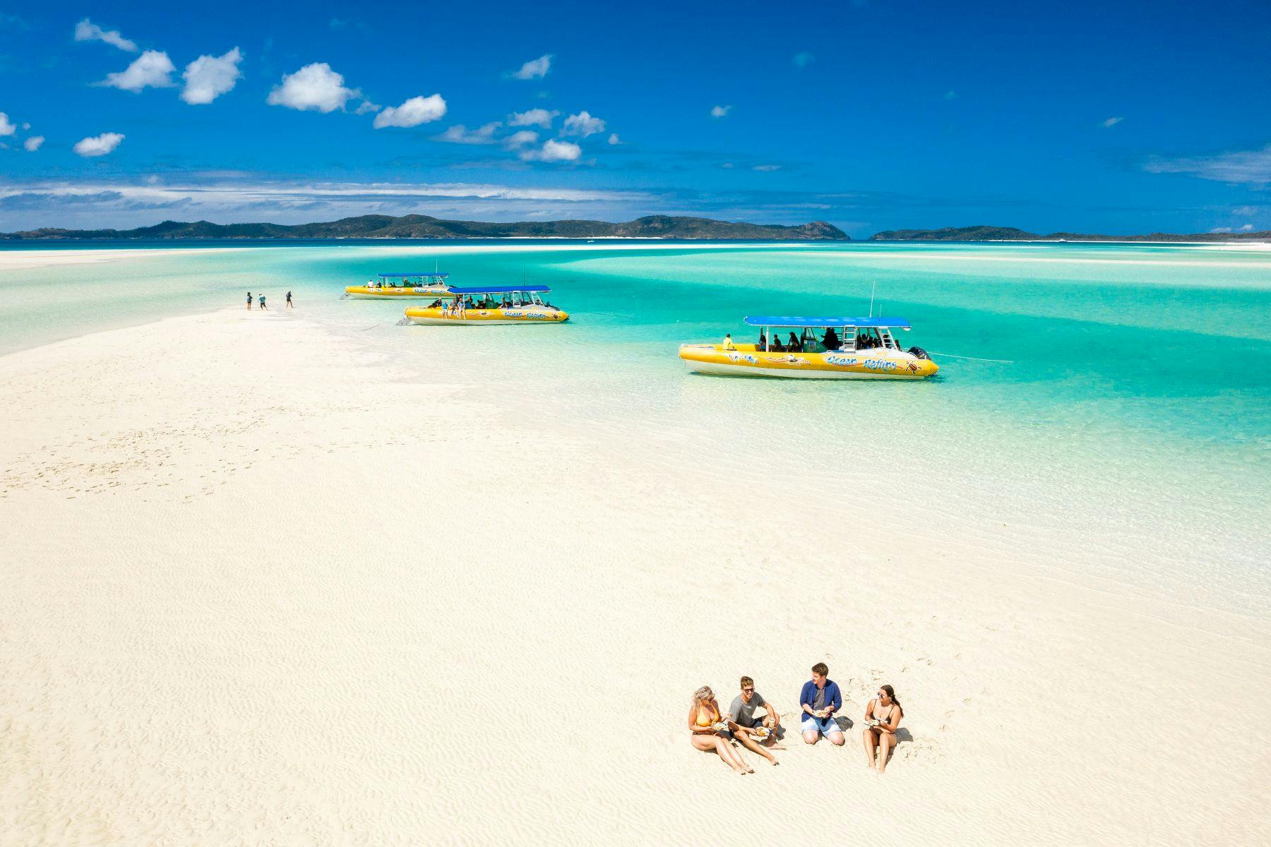 A sunny beach with people sitting on white sand and yellow boats floating in turquoise water in the background.