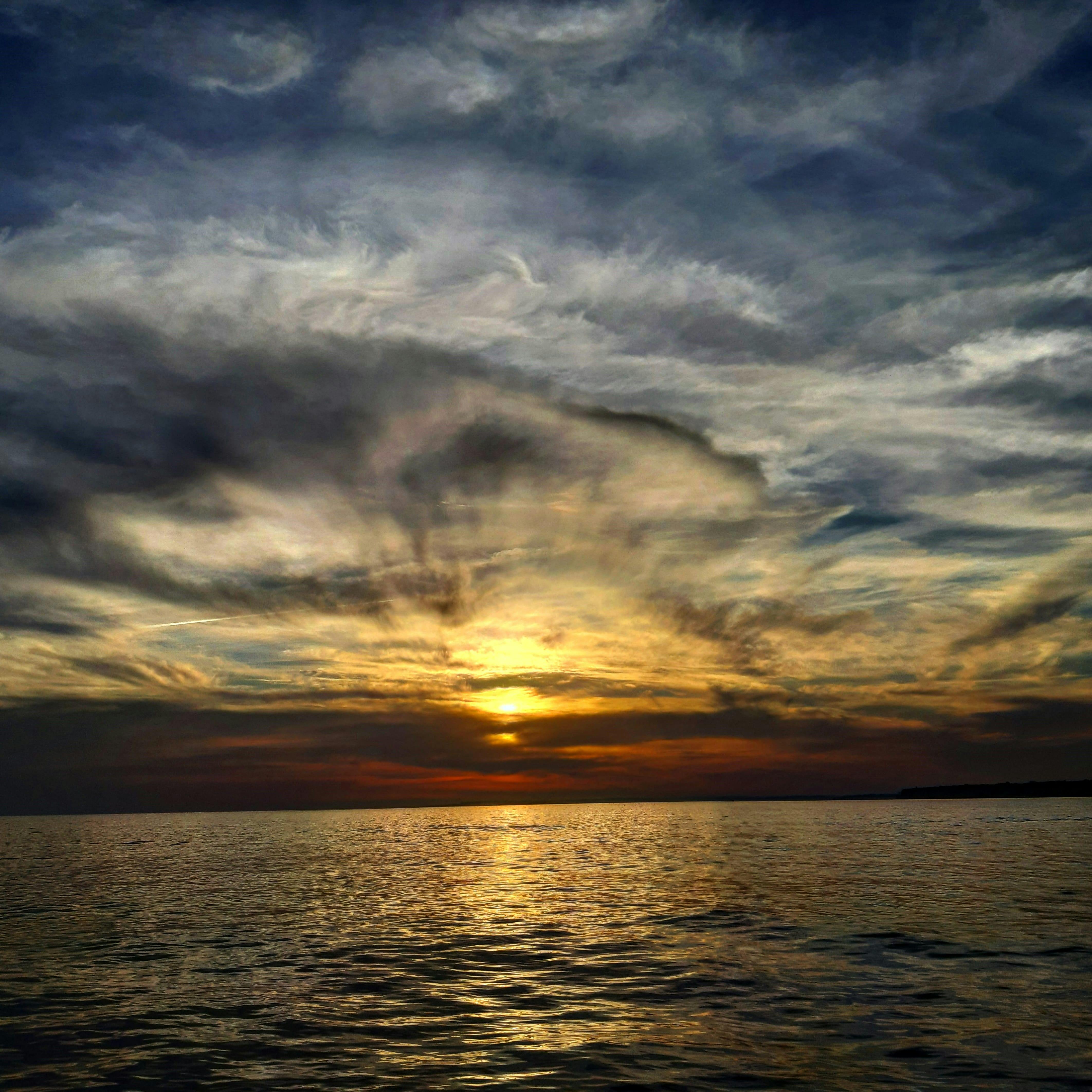 Sunset over calm ocean waters, with the sky filled with dramatic, swirling clouds.
