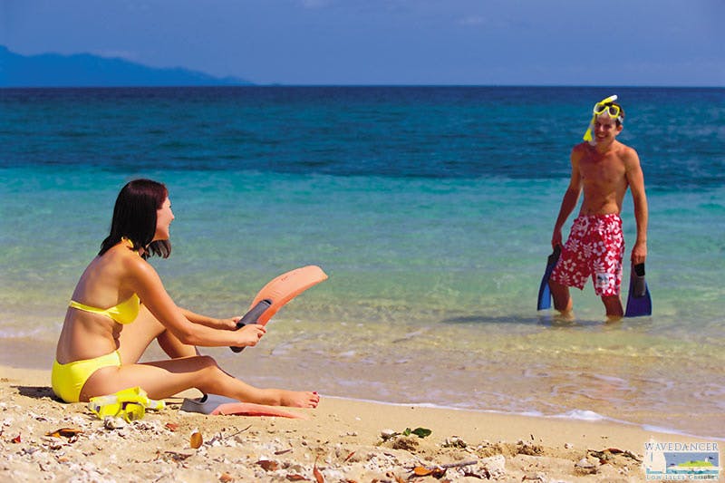 A woman in a yellow bikini sitting on a sandy beach, holding orange snorkeling fins, with clear blue sea and distant mountains.