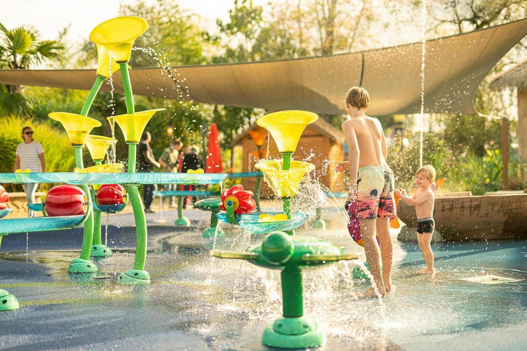 A colorful water play area with yellow funnels, spraying water, and children playing, surrounded by greenery and shade structures.