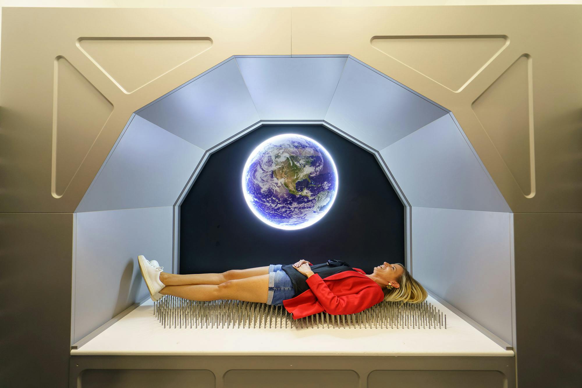 A woman in a red jacket lies on a bed of nails in a futuristic room with an illuminated Earth image on the wall behind her.