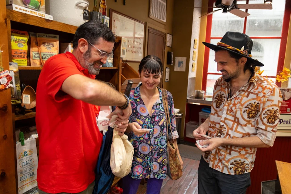 Three people stand together smiling inside a cozy shop. One man in a red shirt, a woman in colorful clothing, and a man in a patterned shirt and hat.