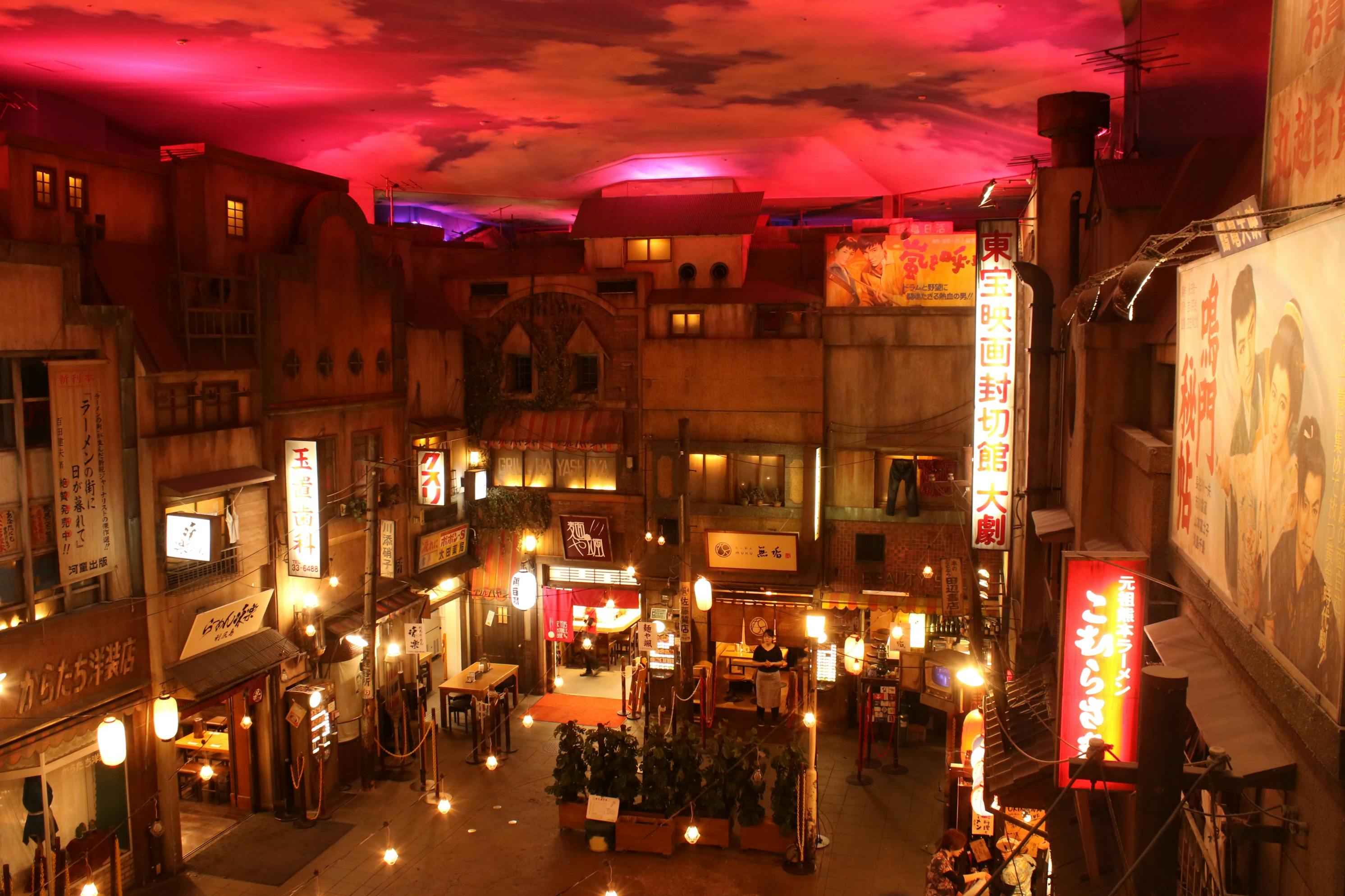 A cozy indoor setting resembling a Japanese street with shops, lanterns, and a vibrant red-lit sky ceiling.