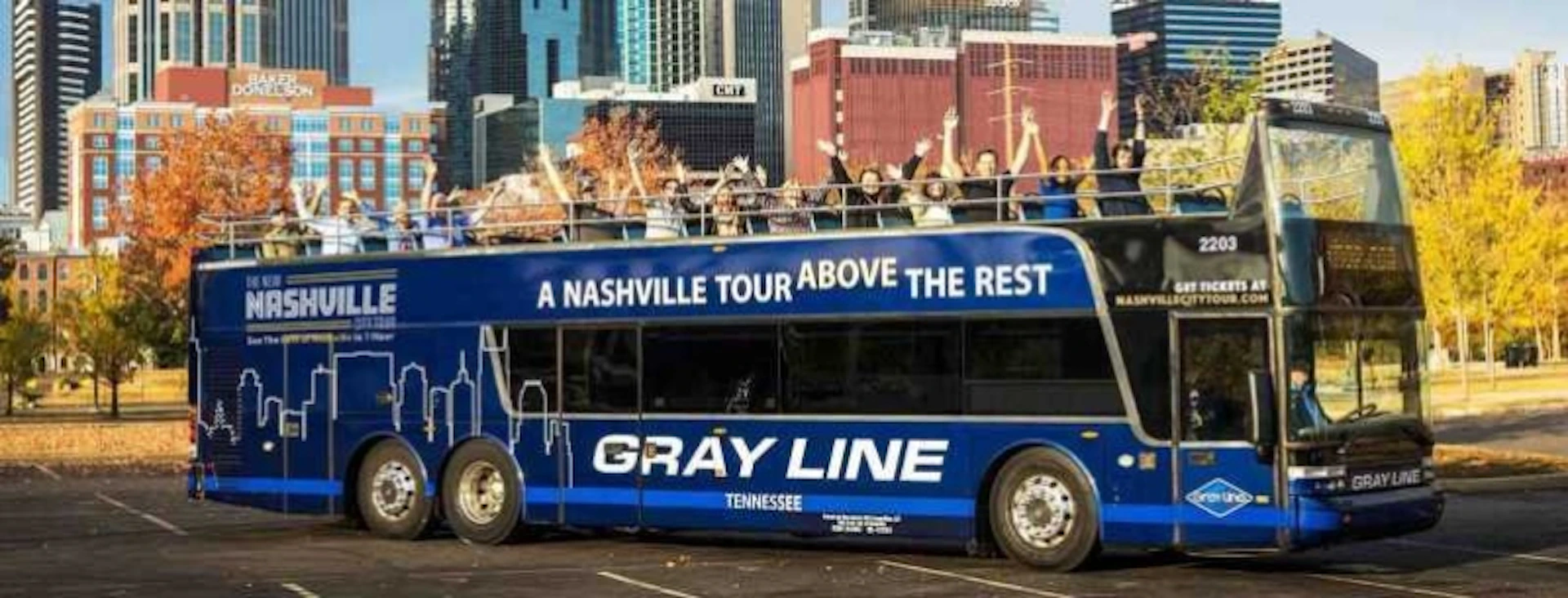 A double-decker tour bus in Nashville with people on the top deck waving. The bus is labeled "GRAY LINE" and "A NASHVILLE TOUR ABOVE THE REST."