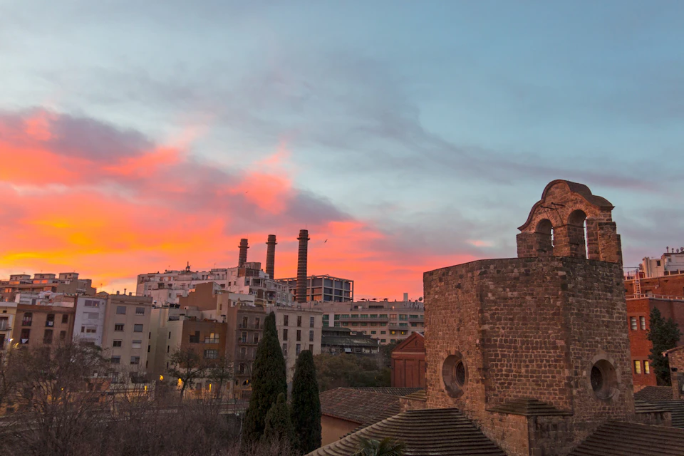 Skyline at dusk with a mix of historic and modern buildings, including three tall smokestacks and a stone structure in the foreground.