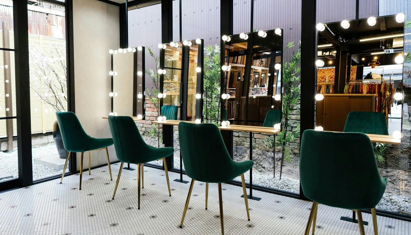 A makeup studio with green chairs and lighted vanity mirrors in front of large windows overlooking an outdoor area.
