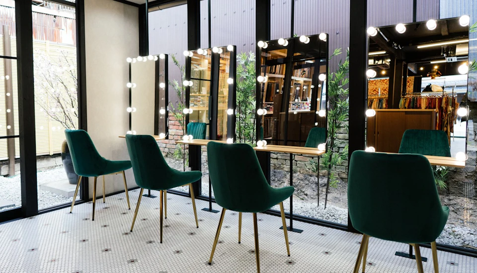 A well-lit makeup station with green chairs and mirrors surrounded by light bulbs, set against a windowed wall with plants outside.