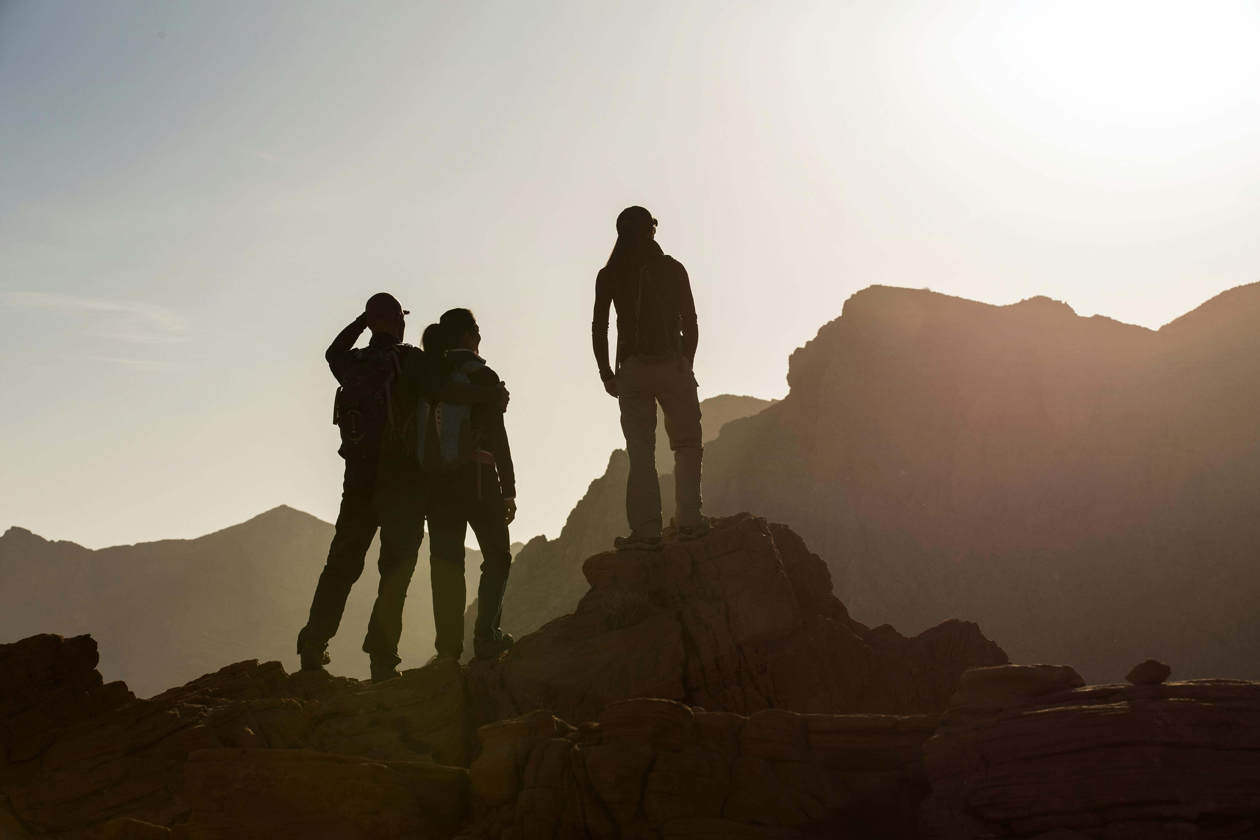 Three people silhouetted against a bright sky stand on a rocky mountain peak with distant mountains in the background.