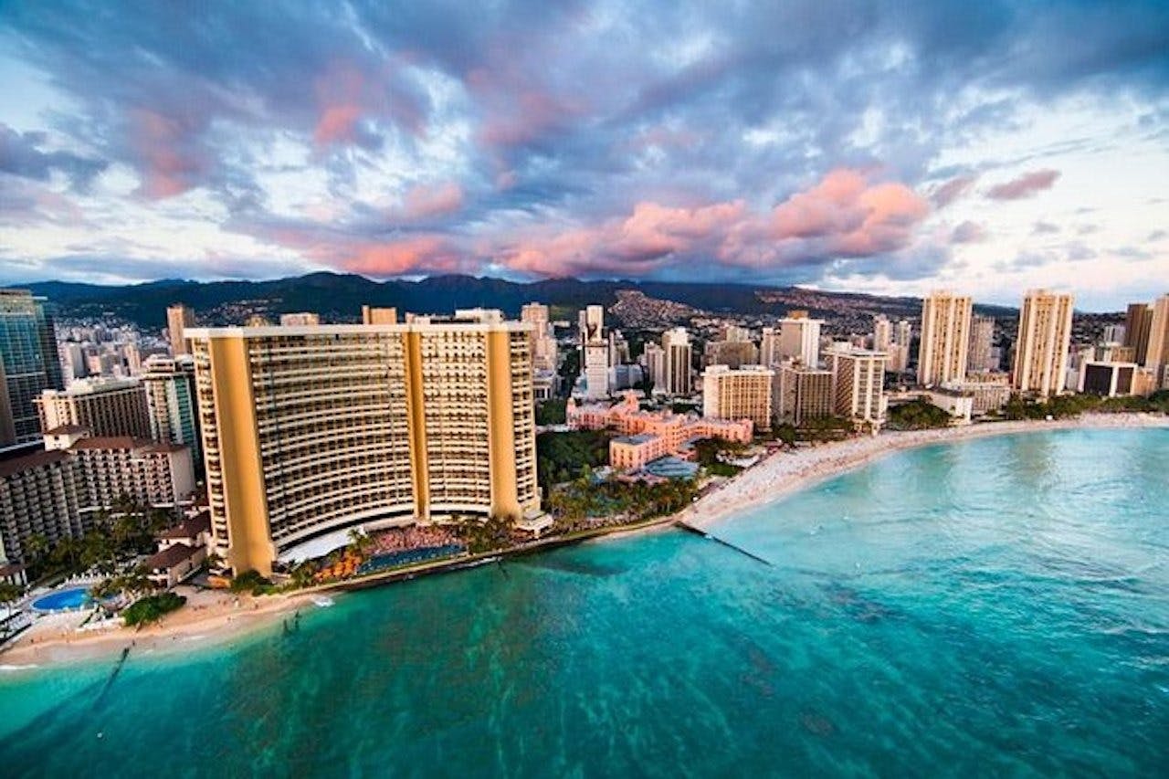 Aerial view of a coastal city with skyscrapers, a sandy beach, and clear blue-green ocean under a partly cloudy sky at sunset.