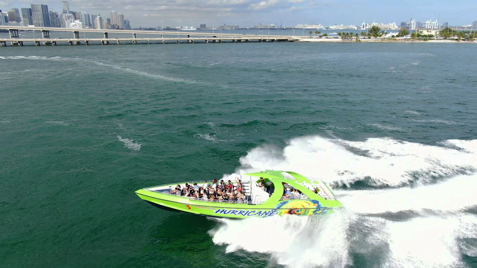 A green and yellow speedboat named "Hurricane" with passengers speeding through the water near a bridge and city skyline.