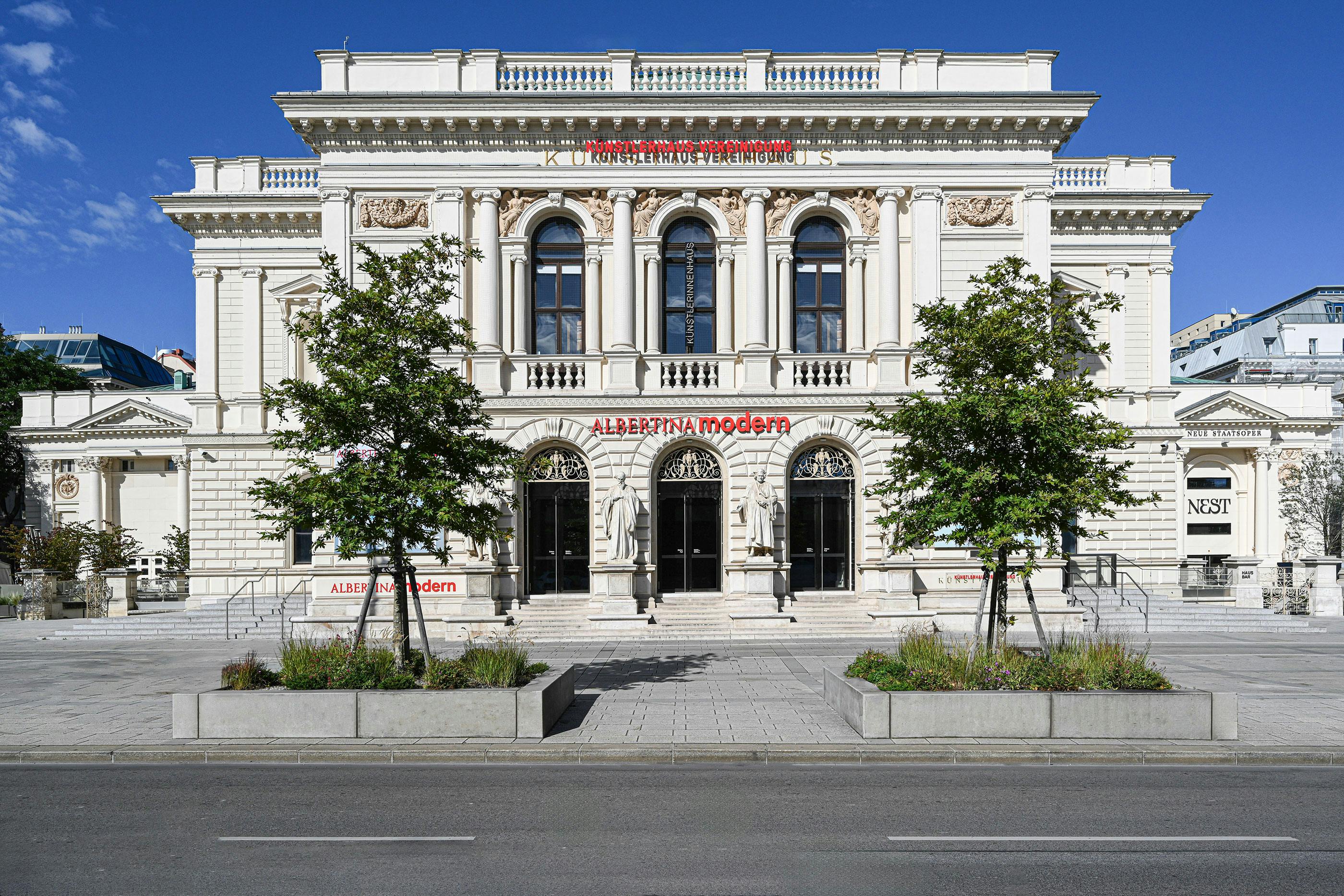 Ornate building with columns and arched windows, labeled &#34;Albertina Modern,&#34; flanked by trees on a sunny day.