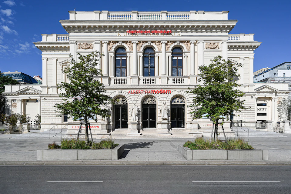 Ornate building with columns and arched windows, labeled "Albertina Modern," flanked by trees on a sunny day.