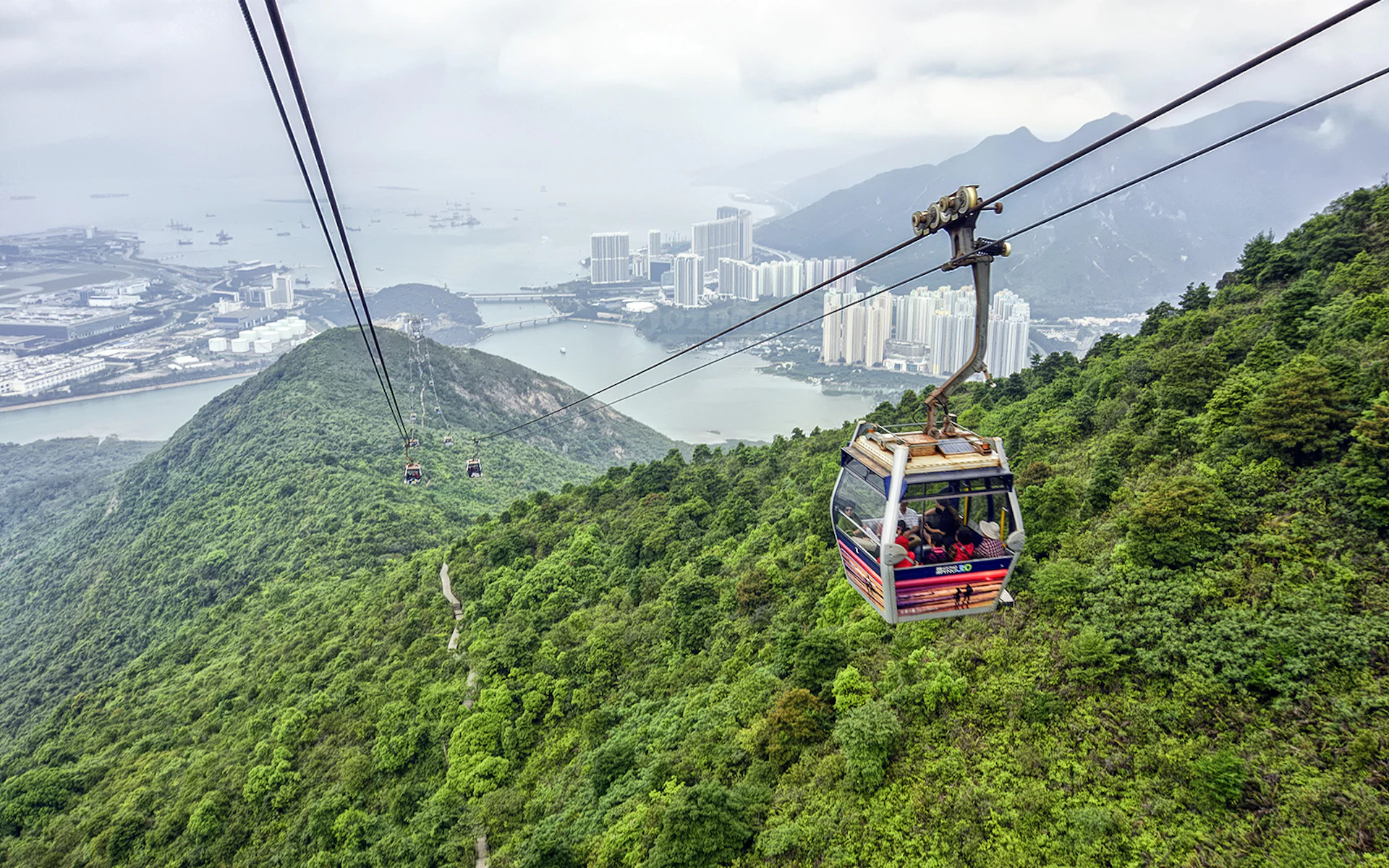 Cable cars moving over a green mountainous landscape, with a city, sea, and distant mountains in the background.