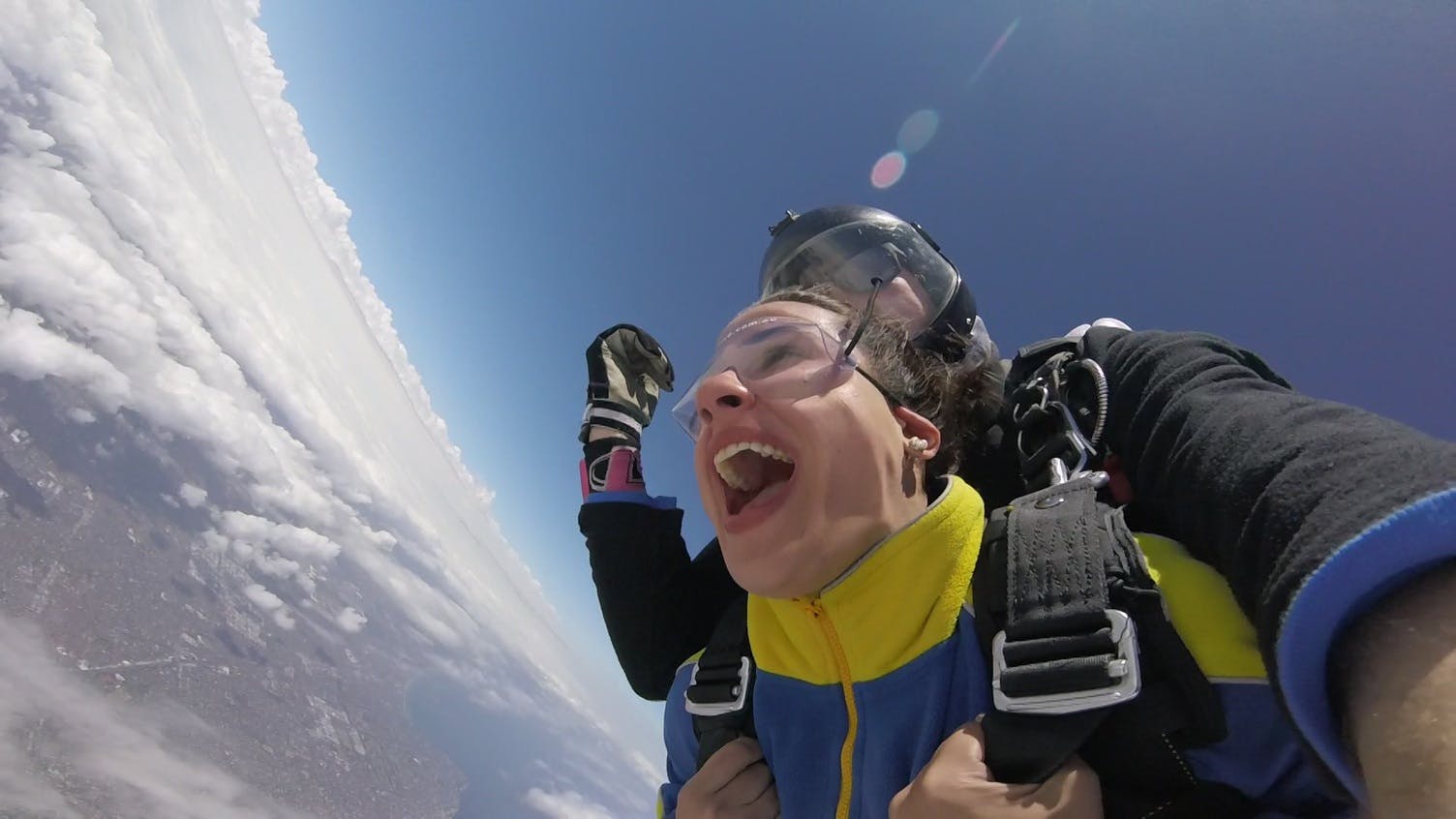 Person skydiving with a wide open mouth, strapped to an instructor against a backdrop of sky and clouds.